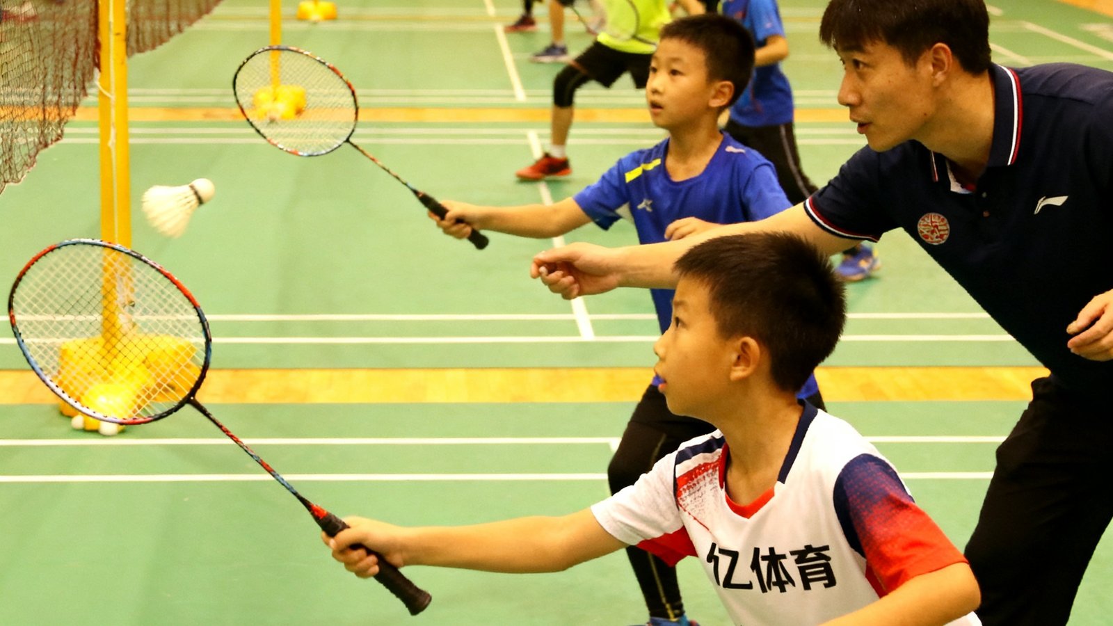 三亿体育青少年基础班学员在教练指导下进行正手高远球挥拍练习，场地内多名小学员有序排列，教练逐一纠正动作。